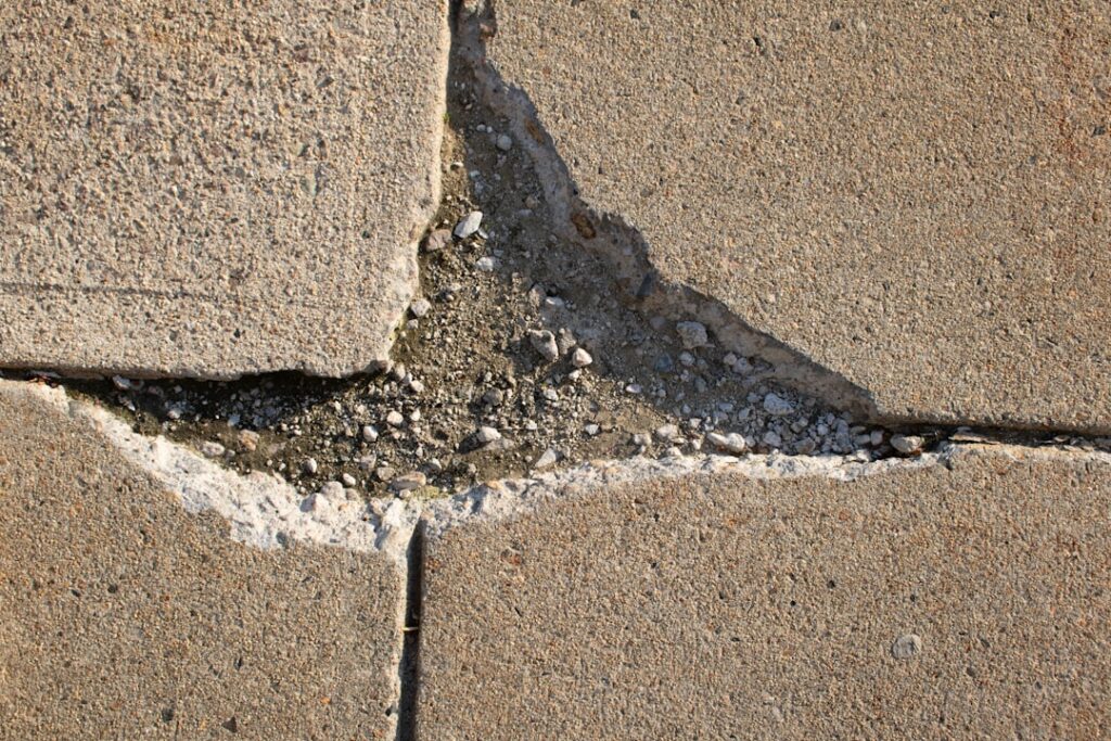 A close-up of cracked, weathered concrete pavement with visible spalling and surface flaking.
